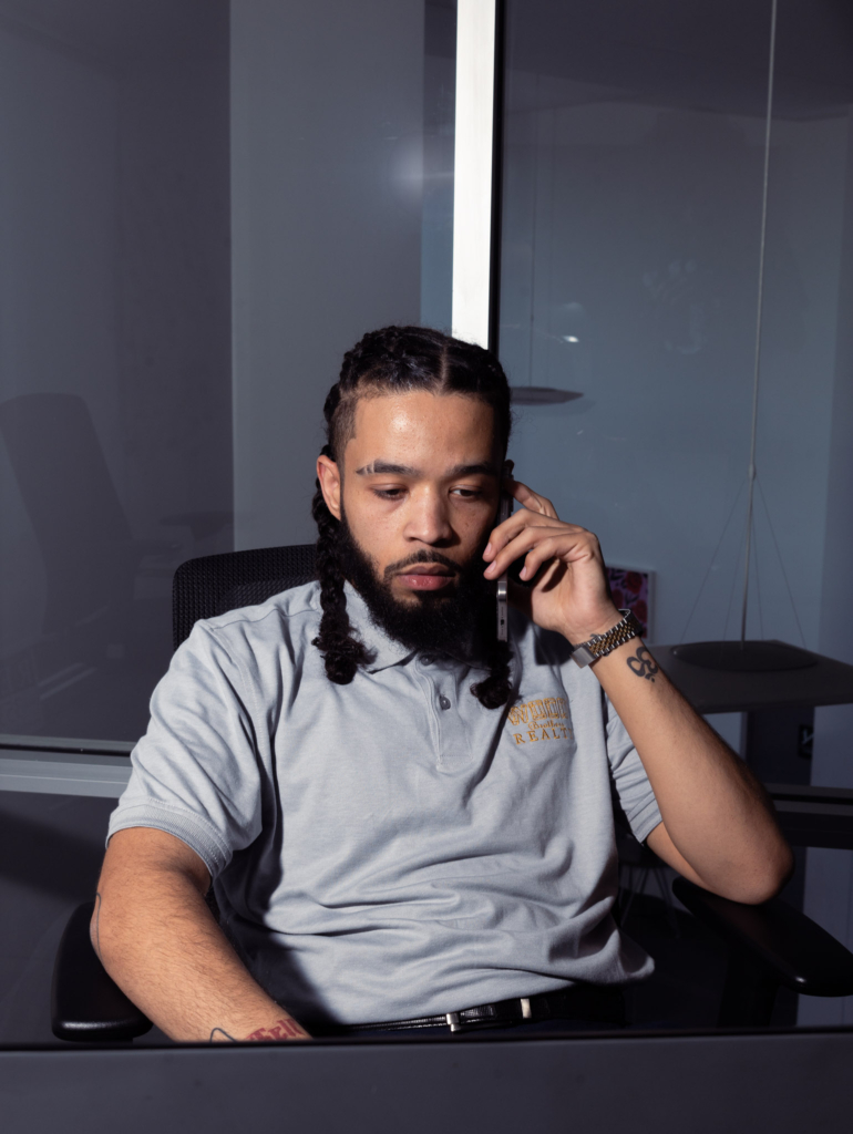 Khalil Henry, a Saint Louis real estate agent with Wood Brothers Realty, on the phone while working at his desk, wearing a gray polo with the company logo, focusing on client communication.