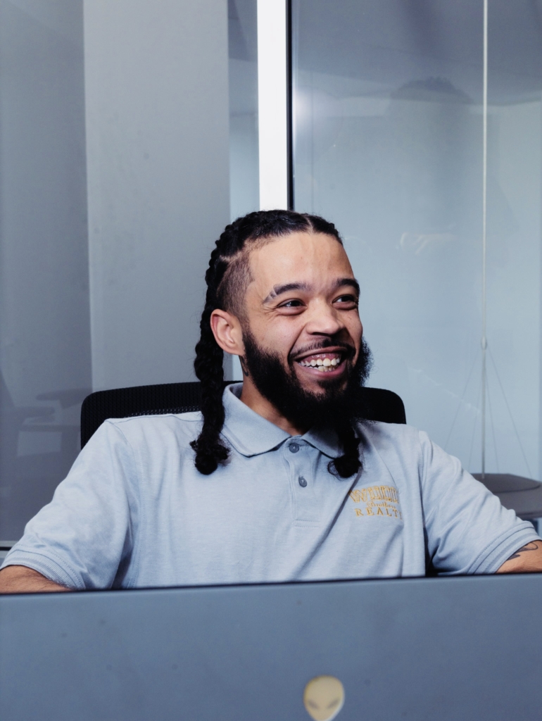 Realtor Khalil Henry smiling in a Wood Brothers Realty polo shirt working at a laptop in an office setting.